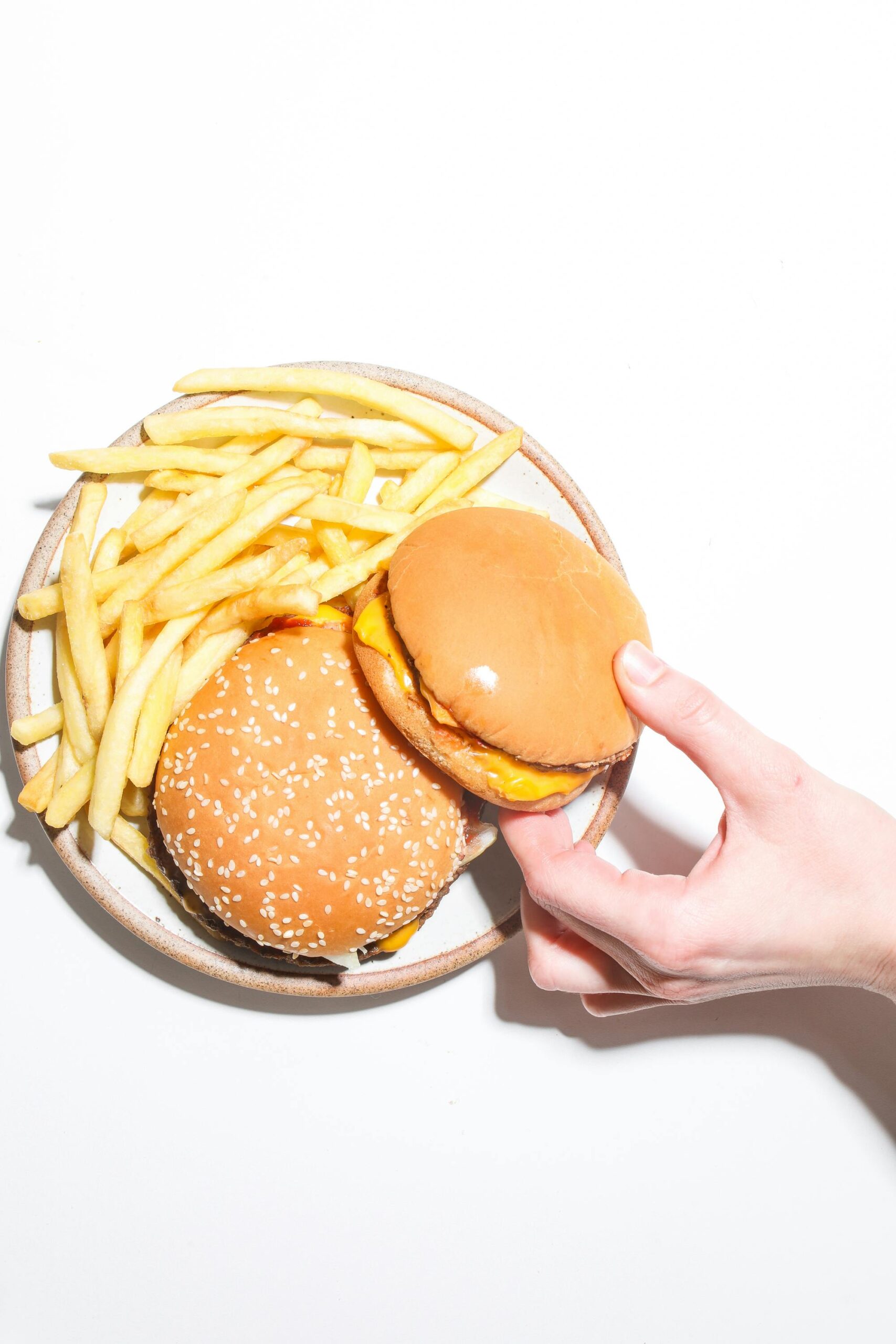 Top view of delicious burgers and crispy fries with a hand reaching for one, on a bright background.